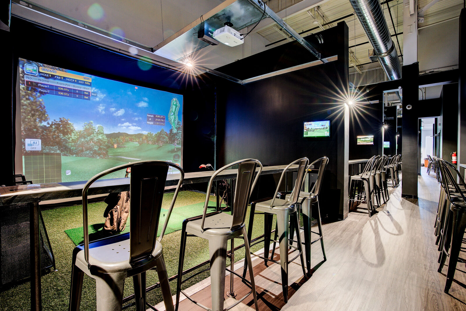 A row of Gamer Golf simulator bays, each with a screen, turf tee box, and bar-height seating.